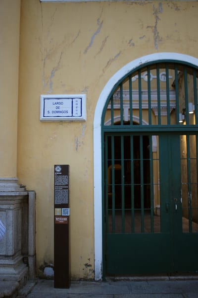 A blue and white tiled street sign on a yellow wall beside a green gate