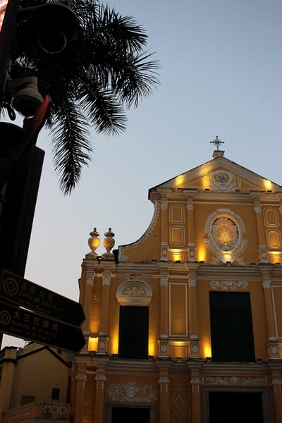 The lit facade of St Dominic's Church in Macau at dusk, a palm frond overhead